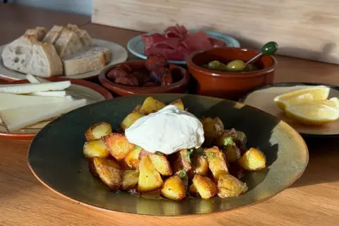 En tallrik med krispiga patatas bravas toppad med en klick krämig sås, omgiven av bröd, ost, chorizo, oliver och citronklyftor.