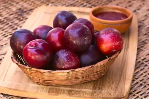 Korb mit dunklen, glänzenden Pflaumen. Vorsicht: unreife Pflaumen giftig! Daneben eine kleine Schale mit Pflaumenmus.