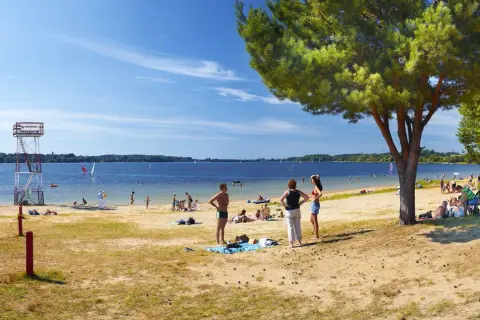 Słoneczna plaża nad jeziorem goczałkowickim. Ludzie odpoczywają, kąpią się i pływają na żaglówkach. Widoczna wieża ratownicza.