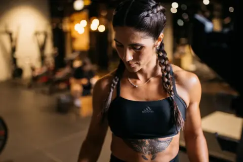 Frau mit definierten Muskeln im Fitnessstudio, trägt Sport-BH und hat Zöpfe.