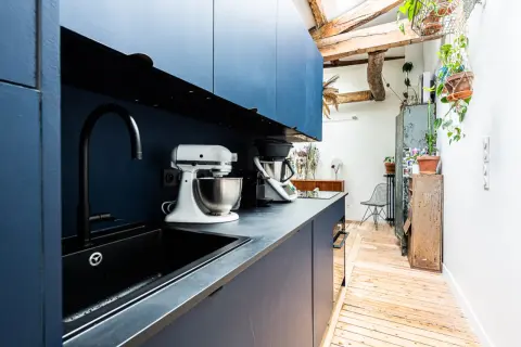 Cuisine moderne avec armoires bleues, évier noir et robots culinaires. L'espace offre une entrée directe dans la cuisine, avec des poutres apparentes et des plantes vertes.