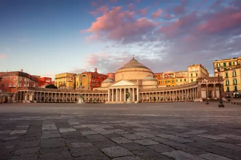 Piazza del Plebiscito a Napoli, con il Palazzo Reale e la Basilica di San Francesco di Paola, sotto un cielo rosato al tramonto.