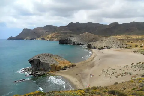 Paisaje de las mejores calas de Cabo de Gata, con formaciones rocosas, arena dorada y mar turquesa bajo un cielo nublado.