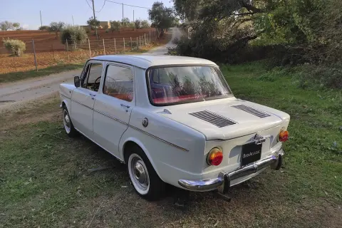Vit Simca-bil parkerad på en gräsbevuxen stig, med olivträd och åkermark i bakgrunden.