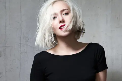 Blonde Frau mit einem geraden Bob und rotem Lippenstift, die provokativ in die Kamera blickt.