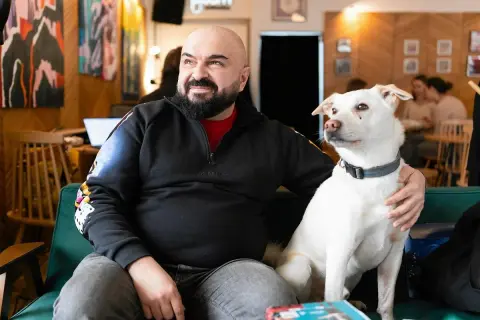 Łysy mężczyzna z brodą siedzi na kanapie z białym, dzikim psem.