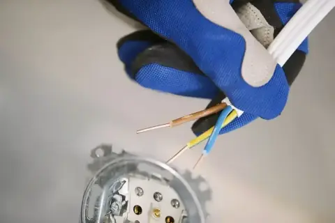 Ręka w rękawicy trzyma przewody elektryczne, przygotowując się do podłączenia fazy w gniazdku. Widoczne są przewody brązowy, niebieski i żółto-zielony.