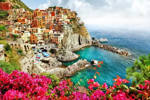 Färgstarka hus klättrar uppför klipporna vid den italienska kusten. Klart turkost vatten och rosa blommor ramar in vyn.