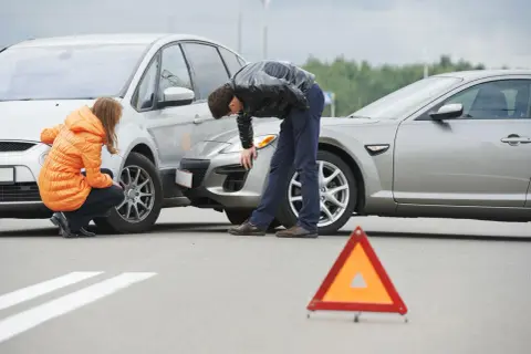 Incidente stradale con due auto danneggiate. Una donna in giacca arancione e un uomo esaminano i veicoli. Triangolo di segnalazione a terra. Nessun segno di omissione di soccorso.