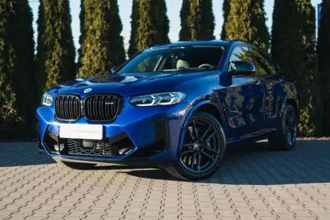 Błękitny suv BMW X4 M Competition błyszczy na tle zielonych drzew.