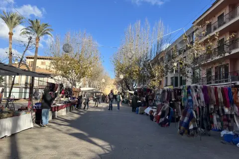 Livlig marknad i Alcudia med stånd som säljer kläder och accessoarer. Palmträd och byggnader kantas av gatan.