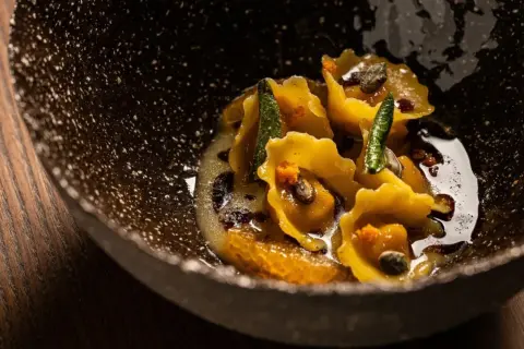 Tortelli di zucca dorati, guarniti con salvia croccante e arancia, in una ciotola scura e rustica.