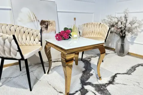 Elegancki stolik do salonu ze złotymi nogami, szklanym blatem, obok krzeseł i wazonu z kwiatami.