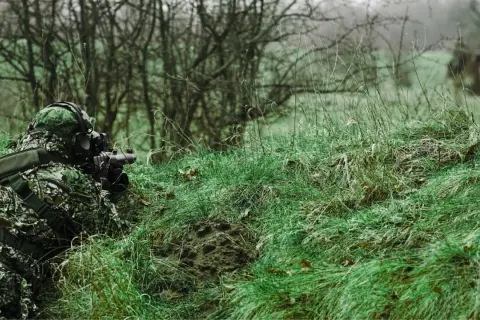 Jugador de airsoft que dispara desde la hierba alta, camuflado y con equipo táctico.