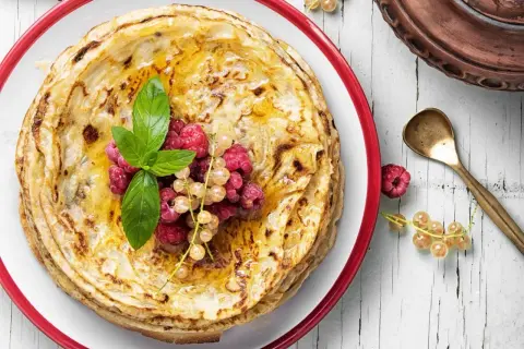 Ein Stapel goldbrauner Pfannkuchen, garniert mit Beeren und Minze. Der Pfannkuchenteig ist perfekt gebacken und mit Honig beträufelt.