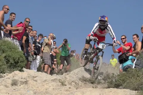 Kolarz w akcji podczas zawodów MTB, otoczony przez widzów i fotografów.