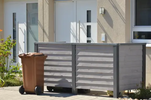 Nowy domek na śmieci to świetny sposób, jak ukryć kosz na śmieci w ogrodzie. Brązowy kosz stoi obok drewnianej obudowy.