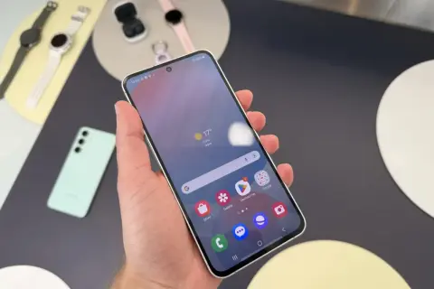Ręka trzyma smartfon Samsung, pokazując ekran z pogodą i ikonami aplikacji. Obok leżą smartwatch i słuchawki. Czas przed ekranem Samsunga to chwila relaksu.