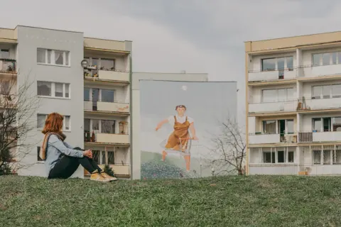 Dziewczyna z rudymi włosami siedzi na trawie, patrząc na mural z kobietą pchającą wózek na tle bloków na gdańskiej Zaspie.