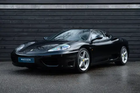 Una splendida auto nera, una Ferrari 360 Modena, parcheggiata su asfalto bagnato davanti a un muro di legno scuro.