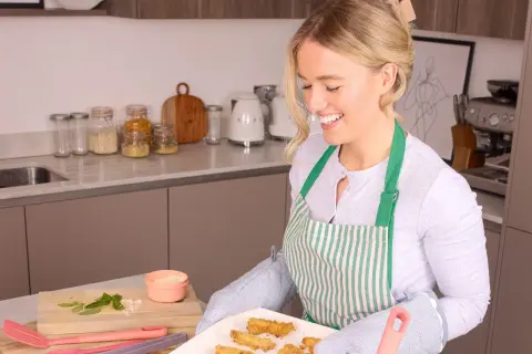 Lächelnde Frau mit Schürze holt gebackene Hähnchenstreifen aus dem Ofen. Perfekt für das Meal Prep!