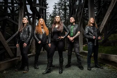 Fünf Musiker in Lederjacken, die an Metal Bands der 80er erinnern, posieren vor einer Holzkonstruktion im Wald.
