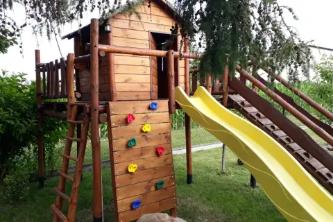 Drewniany plac zabaw z domkiem, zjeżdżalnią, ścianką wspinaczkową i drabinką.