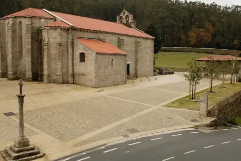 El monasterio de Soandres, con su piedra gris y tejados rojos, se alza majestuoso ante un bosque frondoso. Un cruceiro de piedra vigila la plaza empedrada.