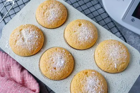 Sechs goldbraune Muffins, mit Puderzucker bestäubt, auf einer hellen Backform. Perfekt für den Thermomix!