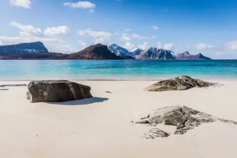 En vit sandstrand vid en turkosblå havsvik, med snöklädda berg i bakgrunden. En idyllisk lofoten beach.