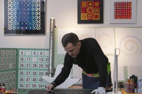 Adriano Attus, artista, disegna cerchi colorati su una tela nera. Sullo sfondo, opere astratte e numeri.