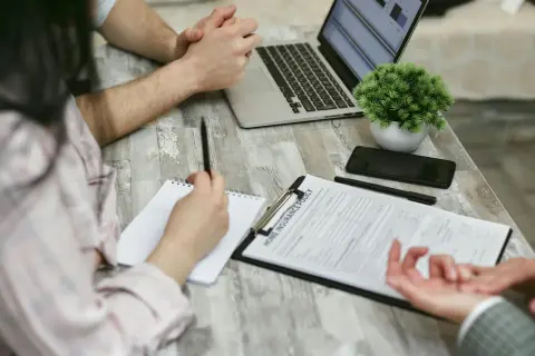 Osoby przy stole omawiają wypowiedzenie umowy ubezpieczenia OC. Na blacie leży laptop, telefon i dokumenty.