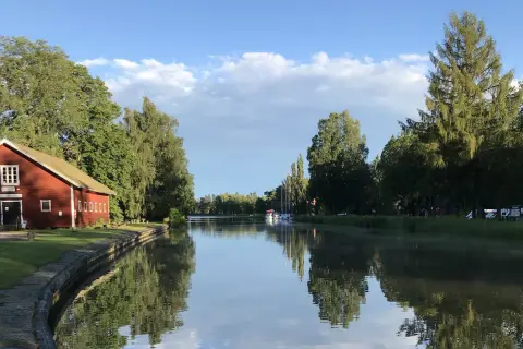 En röd byggnad vid en kanal, troligen Hajstorps slusskafé, med båtar och träd längs vattnet under en blå himmel.