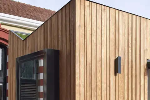 Detalle de una fachada ventilada de madera que contrasta con una pared antigua de ladrillo y piedra.