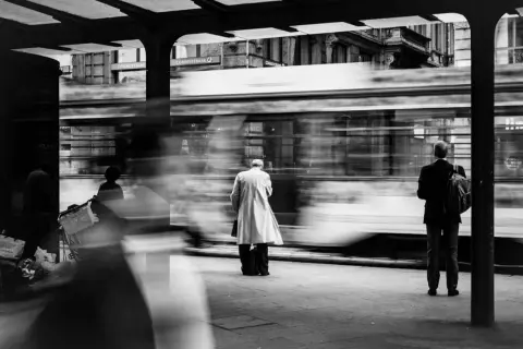 foto di strada: un uomo in impermeabile attende il tram, sfocato dal movimento. Altri passeggeri e la banca Commerzbank sullo sfondo.
