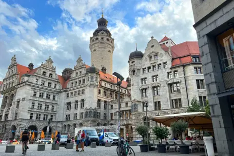 Lipsk trasa: imponujące budynki z wieżą, plac z ludźmi, rowerzystką i kawiarnią pod parasolami.
