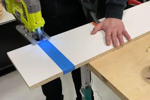 Une scie sauteuse coupe une planche de stratifié, guidée par du ruban bleu. La main d'un bricoleur stabilise la pièce.
