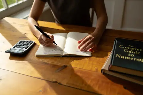 Une personne écrit dans un carnet, une calculatrice et un livre de numérologie à proximité. Cherche-t-elle à trouver une date de naissance gratuite ?
