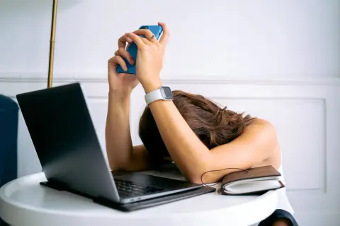 Kobieta z opuszczoną głową na biurku, trzymająca telefon. Wokół laptop i notatnik. Czuje, że ma dość wszystkiego, cytaty z życia.