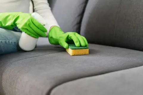 Ręce w zielonych rękawiczkach czyszczą plamy na kanapie gąbką i środkiem w sprayu.