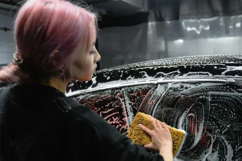 Kobieta z różowymi włosami myje auto gąbką. Czy można myć samochód płynem do naczyń? Piana na karoserii sugeruje eksperyment.
