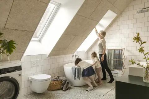 Salle de bain sous pente avec wc suspendu. Une femme et deux enfants admirent la lumière entrant par la fenêtre de toit.