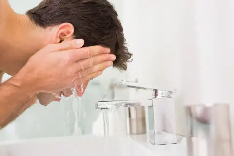 Mann wäscht sein Gesicht mit kaltem Wasser. Er hält seine Hände unter den Wasserhahn, um das Gesicht zu erfrischen.