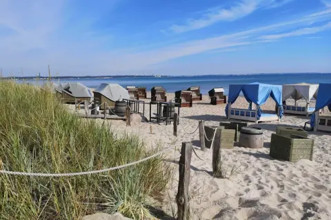 Scharbeutz Strand: Strandkörbe, Liegen unter blauen und weißen Zelten, Tische und Fässer am Sandstrand.