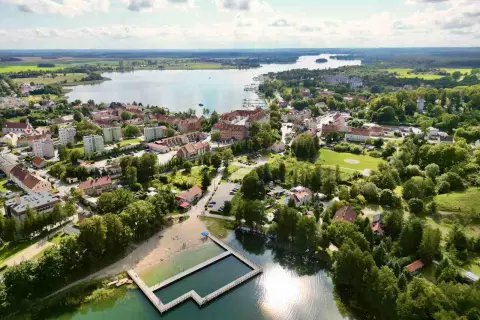 Widok na Ryn z lotu ptaka. Widać jezioro, miasto, plażę i basen. To świetne miejsce na wakacje, pełne atrakcji dla dzieci.