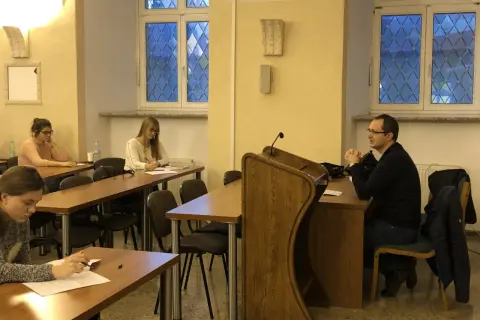 Referendarz sądowy prowadzi szkolenie. Kilka osób siedzi przy ławkach, słuchając i notując.