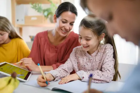 Uśmiechnięta mama pomaga dzieciom w nauce. Jedna córka pisze, druga korzysta z tabletu. To przykład wspólnego uczenia się, gdzie powtarzanie słów, jak w epifora, pomaga zapamiętać.