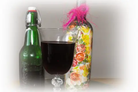 Szklanka z ciemnym winem z wiśni obok zielonej butelki i ozdobionej kwiatami butelki z różową kokardką.