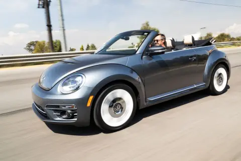 Szary kabriolet Volkswagen Beetle jedzie po drodze. Mężczyzna w okularach przeciwsłonecznych prowadzi auto. To idealny pojazd na letnie wycieczki, jak ta opisana w umowie użyczenia samochodu.