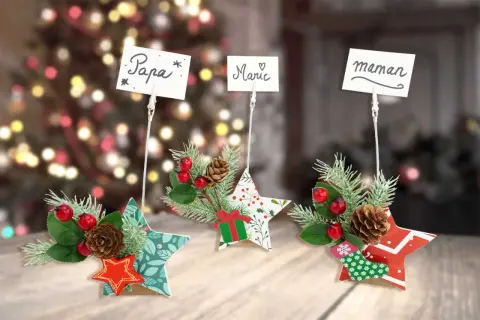 Porte nom original à faire soi-même : étoiles décorées de branches de sapin, baies rouges et pommes de pin, avec des noms écrits sur des cartes.
